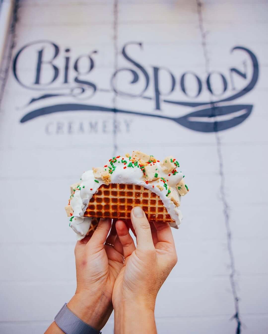 Hands holding a waffle taco filled with ice cream and sprinkles in front of the Big Spoon Creamery sign in Avondale.