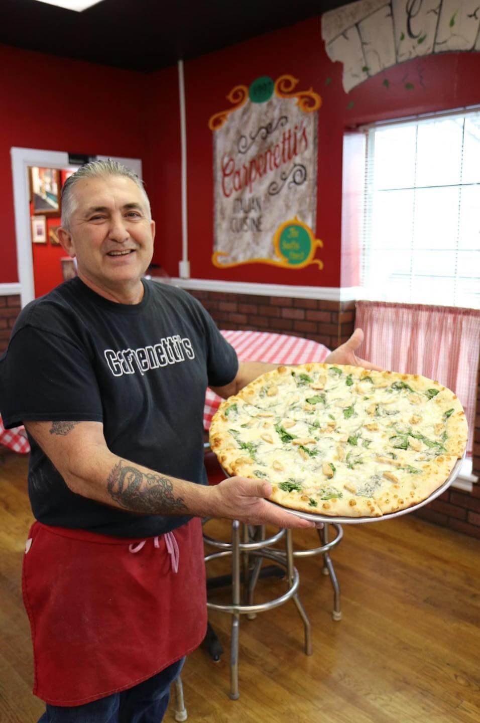 Frank Carpenetti holds a large pizza topped with greens and cheese, standing inside Carpenetti's pizzeria. 