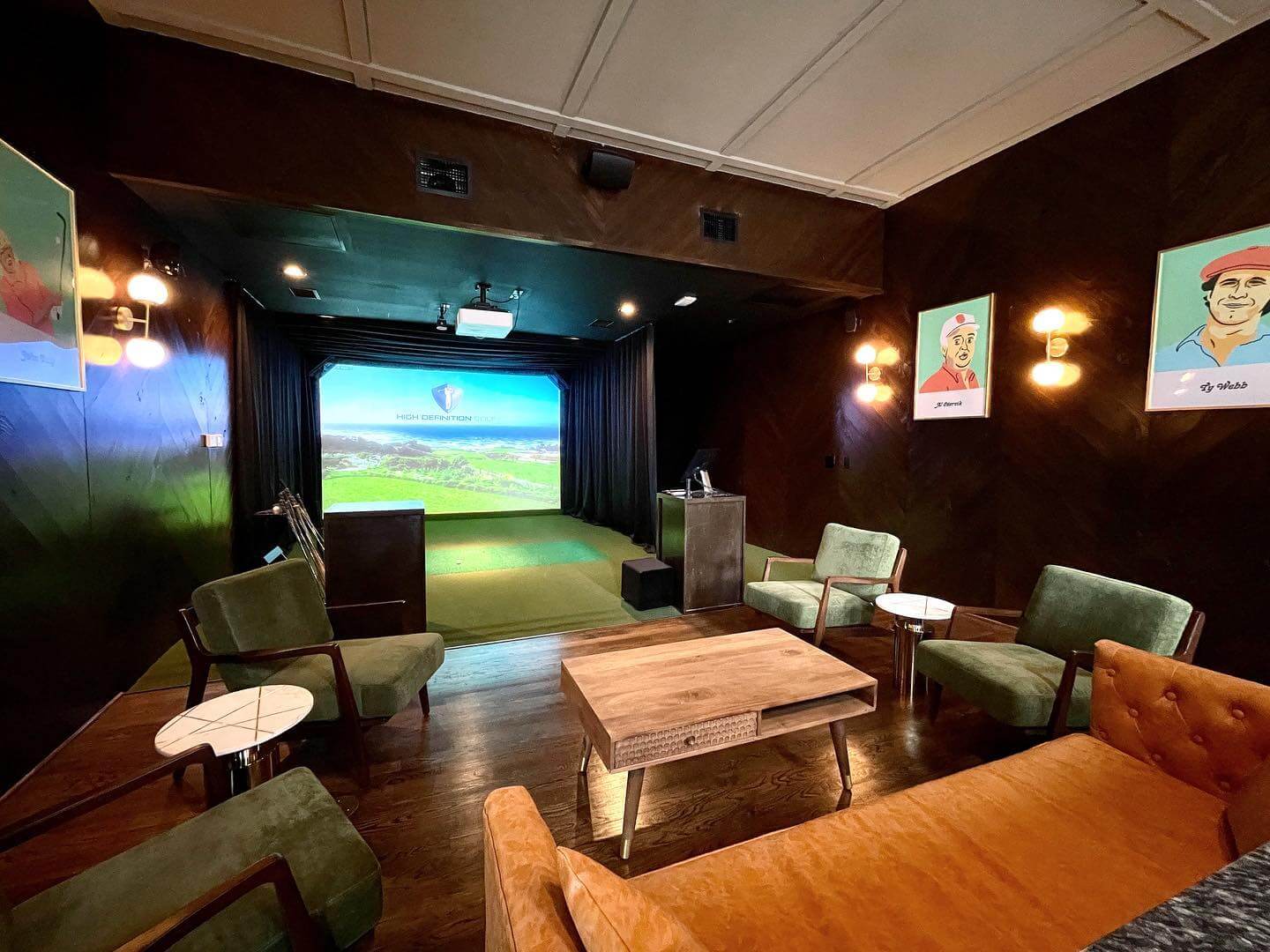 A room with a golf simulator setup, including a screen and green turf along with several green armchairs, two small tables, and a wooden coffee table are arranged around the space at Cinderella Story.