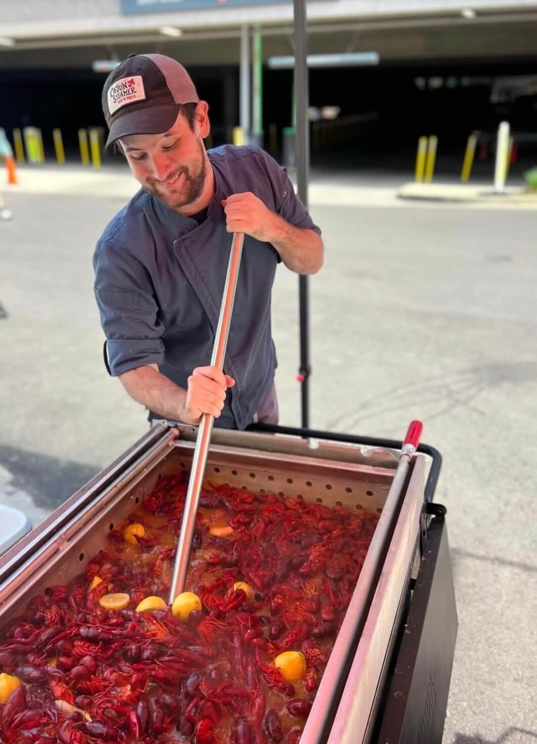 A chef in a cap and apron expertly stirs a large pot of crawfish with lemons outdoors, creating a delicious aroma.