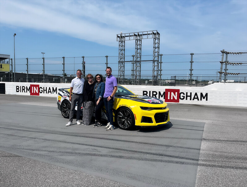 Four people stand in front of a yellow race car on the track, with a bold "BHAM" sign proudly displayed in the background.