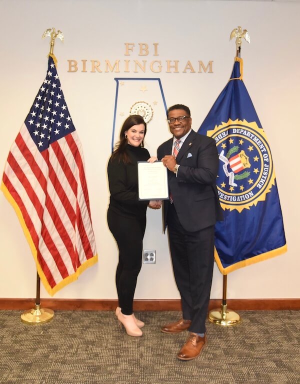 Two people stand in front of American and FBI flags at the BHAM office, holding a document and smiling.