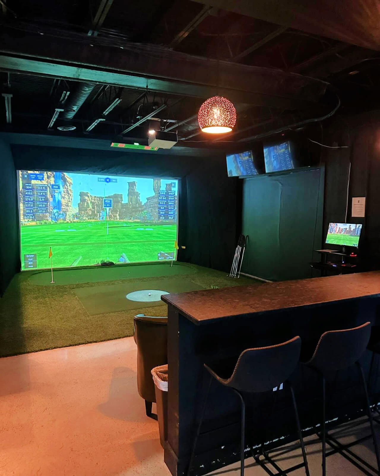 Experience the ultimate indoor golf simulator setup in Birmingham, featuring a screen that brings the fairway to life. With artificial turf, top-notch golf clubs, and two TV screens above a cozy counter with chairs, it's perfect for enjoying immersive golf games anytime.