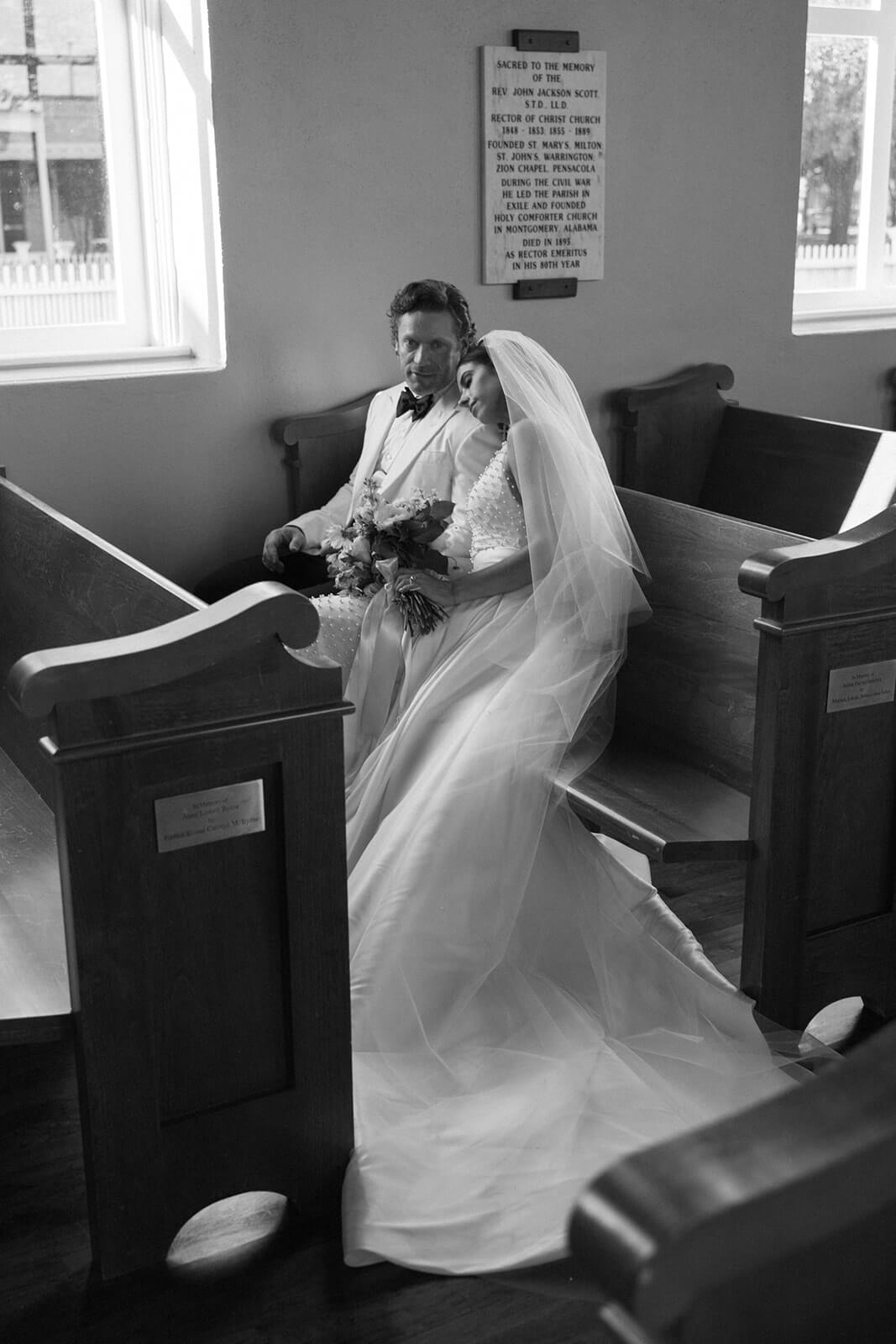 Bride and groom sit together on a church pew in Downtown Historic Pensacola, with the bride resting her head on the groom's shoulder, both adorned in elegant wedding attire.