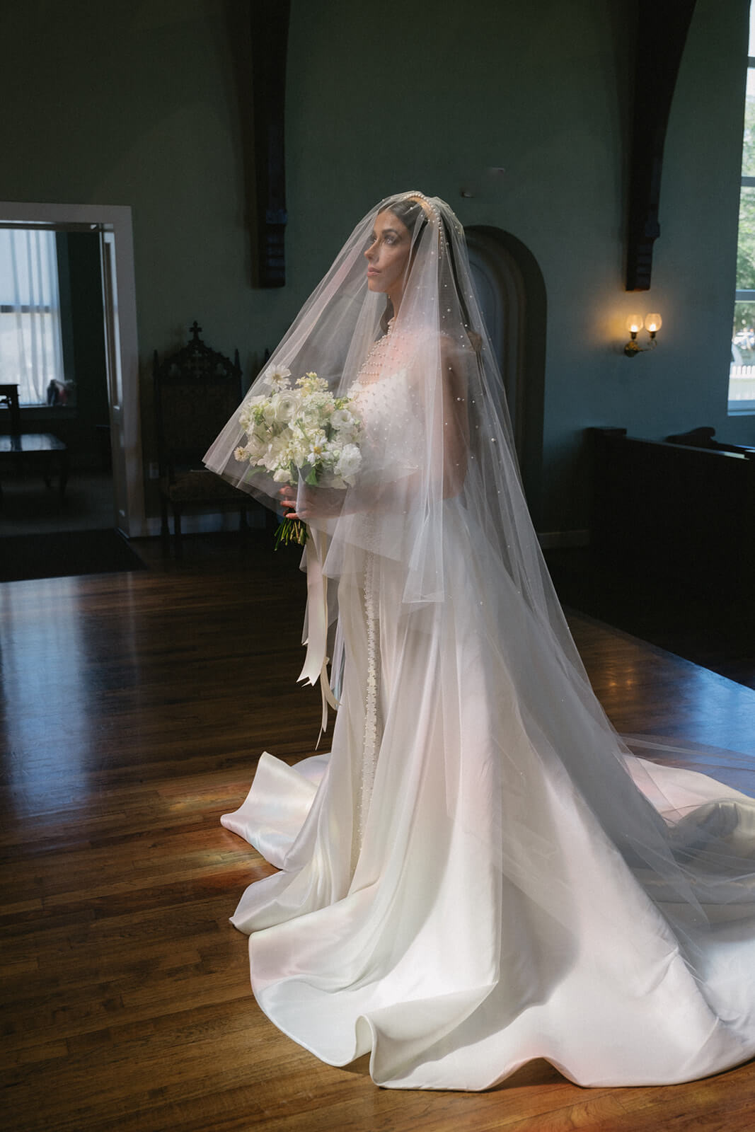 In an elegant indoor setting reminiscent of an Old-World Italian Wedding, a bride in a white gown and veil stands gracefully, her bouquet glowing as sunlight streams through the windows.