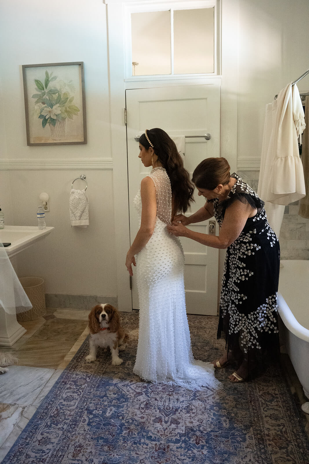 In a charming bathroom reminiscent of Old-World Italian weddings, a woman in a white dress is being assisted by another. A dog sits calmly on the rug nearby, adding to the serene scene.