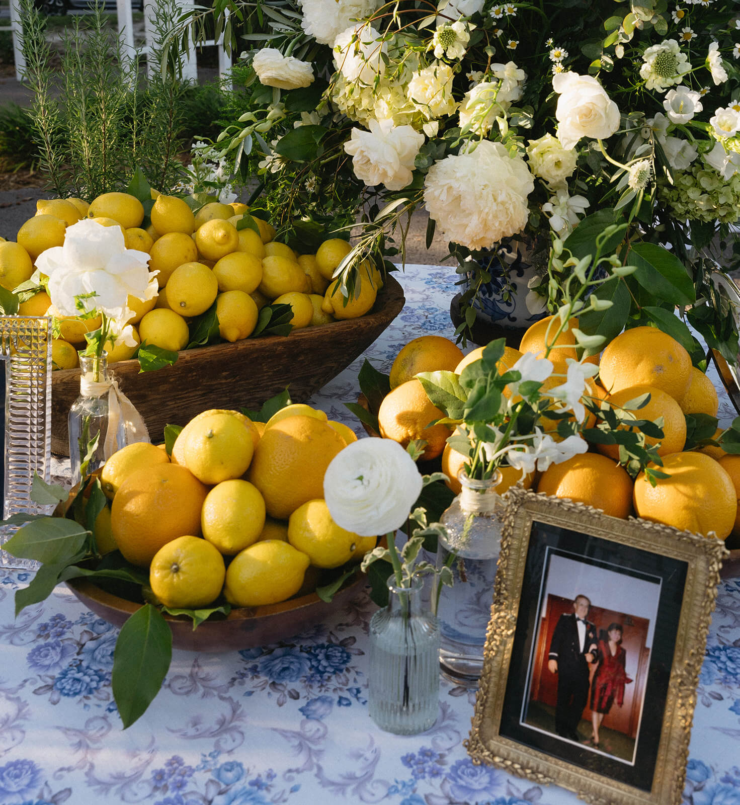 A table adorned with lemons, oranges, flowers, and a framed photo rests on a floral tablecloth, evoking the charm of an Old-World Italian wedding.