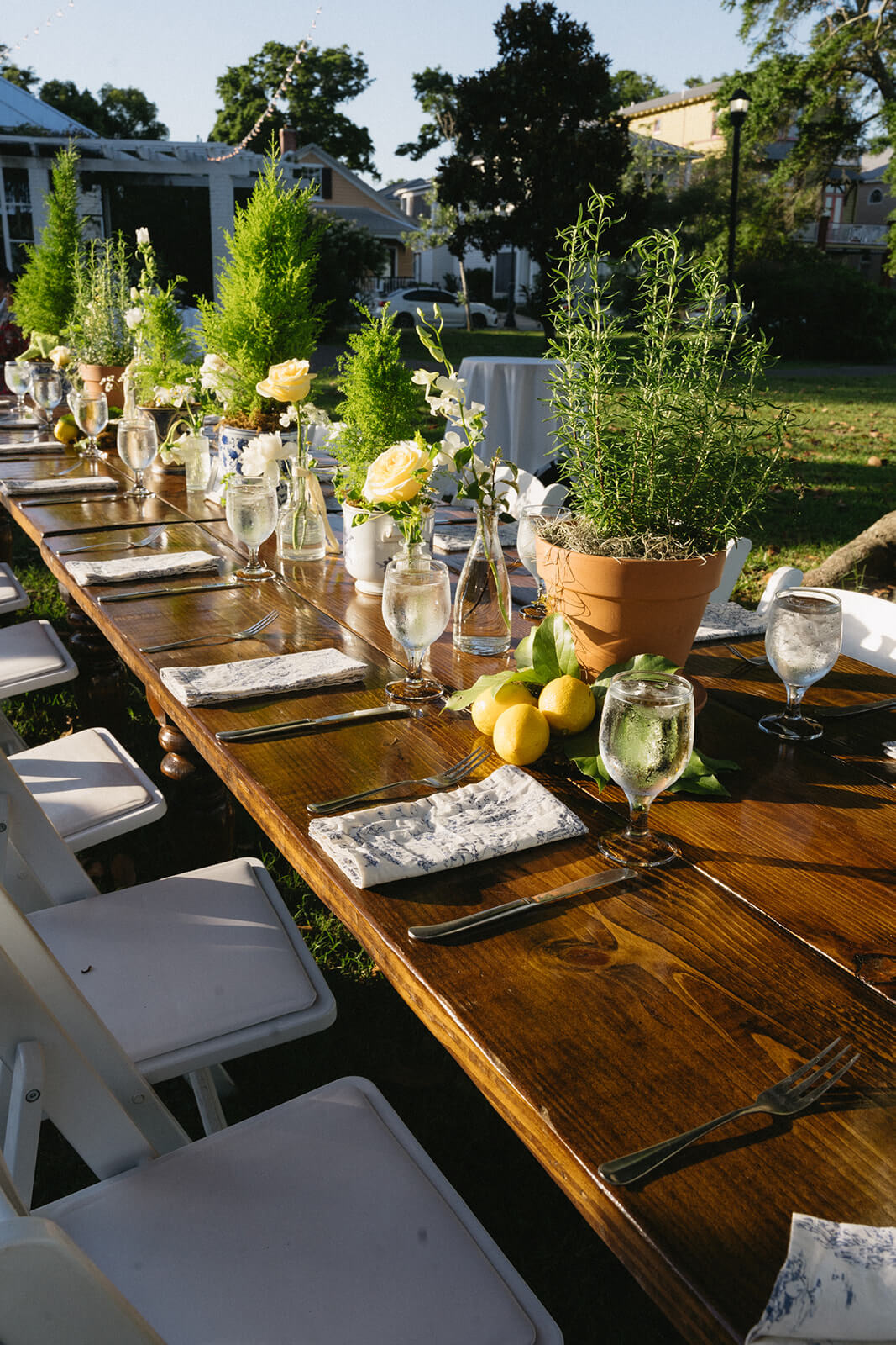 The long wooden table, perfect for an Italian wedding, is set for outdoor dining, adorned with potted plants, lemons, wine glasses, napkins, and cutlery. White chairs surround the table amidst a lush green garden setting.