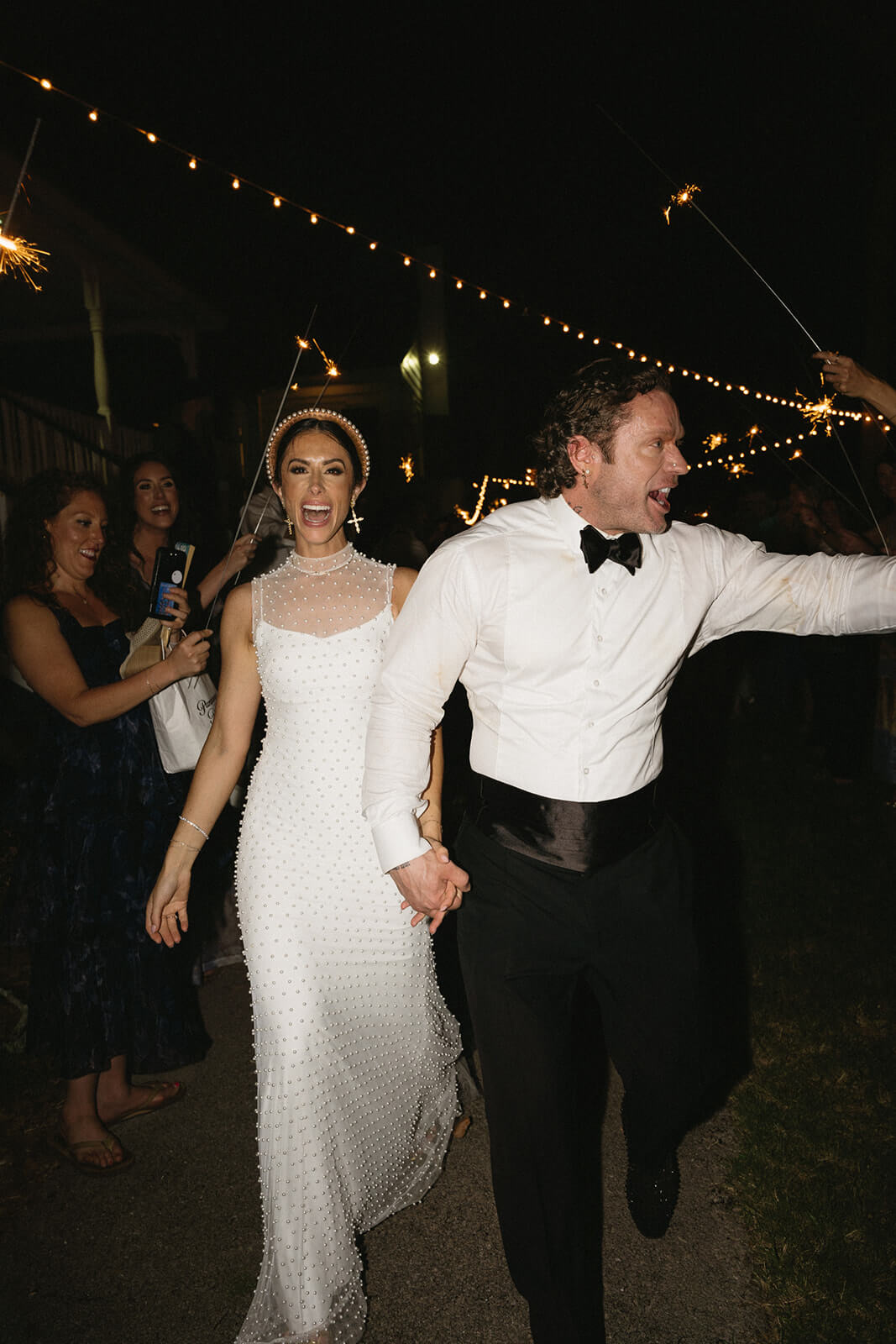 A Birmingham couple in formal attire walks hand-in-hand at night, surrounded by guests holding sparklers.