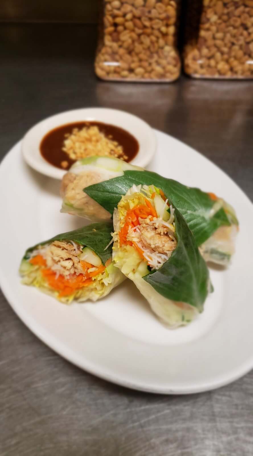 Basil spring rolls filled with vegetables and shredded meat are sliced in half and arranged on a white plate. A dipping sauce with crushed peanuts is in the background from Kati's Thai Cuisine.