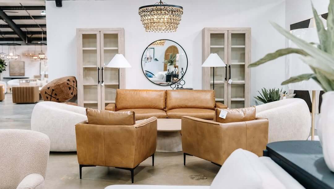A beautiful modern designer living room set featuring a brown leather sofa, two matching armchairs, elegant white curved sofas, a round mirror, stylish cabinets, and a stunning chandelier is pictured in a showroom at Sara Sells.