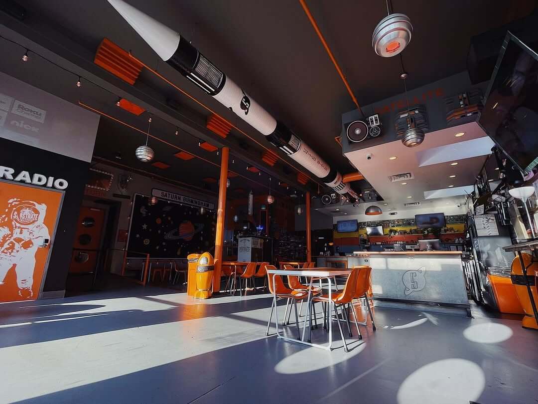 Interior of Avondale's space-themed Satellite cafe with rocket decor, orange chairs, and tables, and a large space rocket replica hanging from the ceiling.