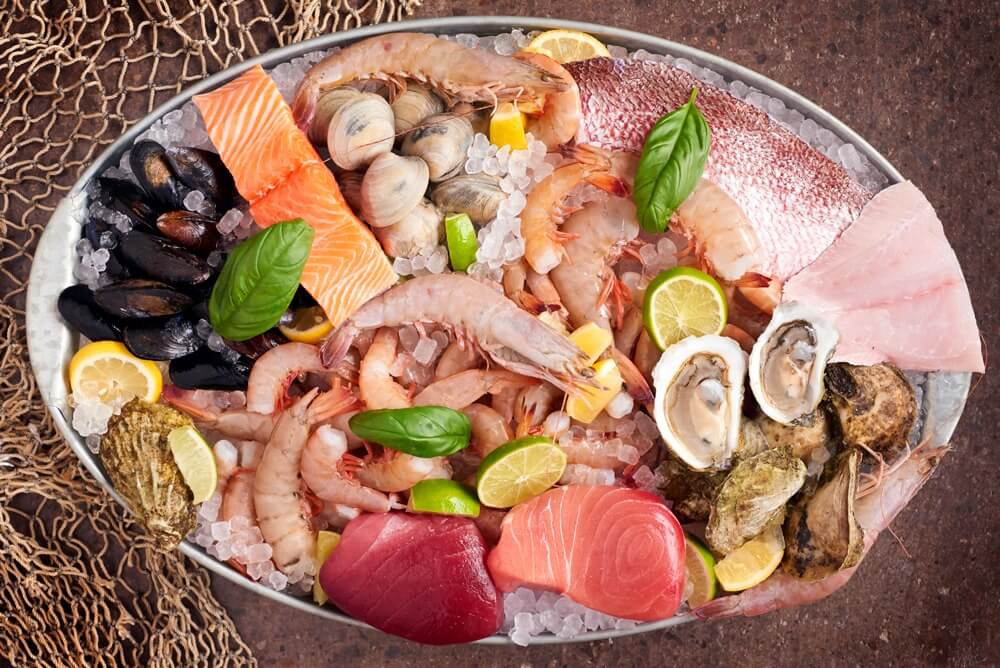 A platter featuring assorted raw seafood delights—shrimp, crawfish, clams, mussels, salmon, tuna, oysters, and a red fish fillet from Snapper Grabber's Land & Sea market, garnished with lemon slices and basil leaves.