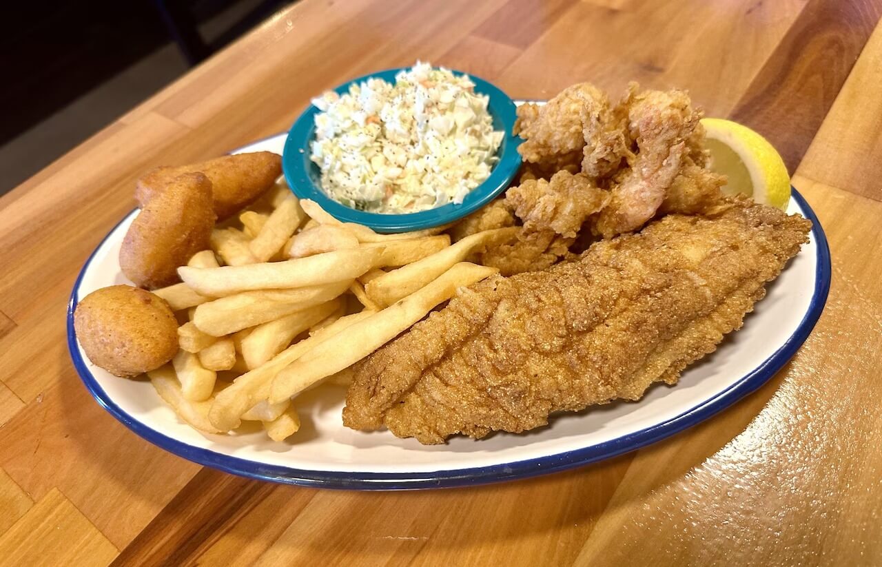 A plate from The Ark featuring fried fish, shrimp, hush puppies, French fries, a lemon wedge, and coleslaw on a wooden table.