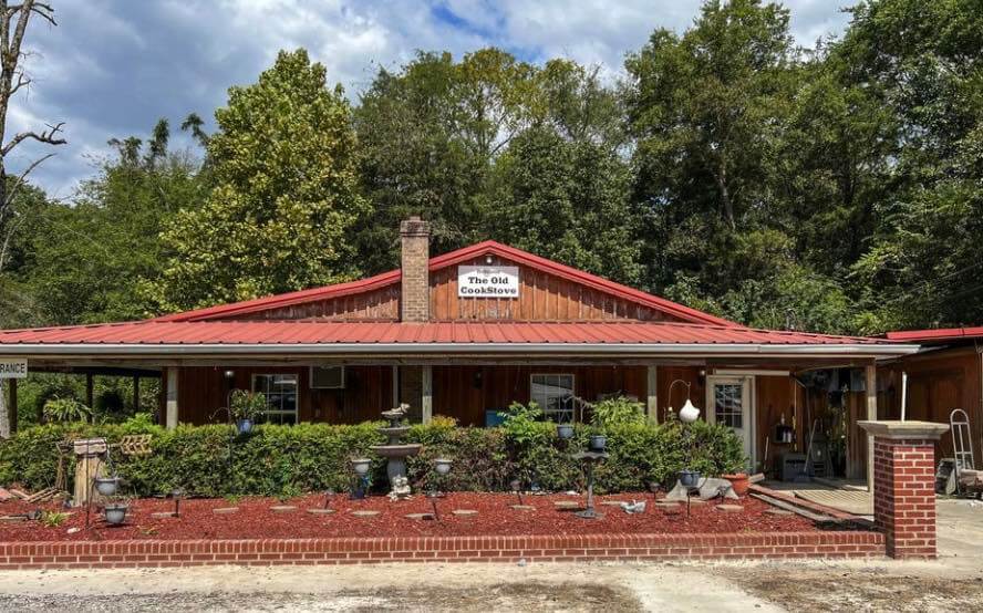A rustic building with a red metal roof, sign reading "The Old Cookstove," surrounded by greenery and garden decorations.