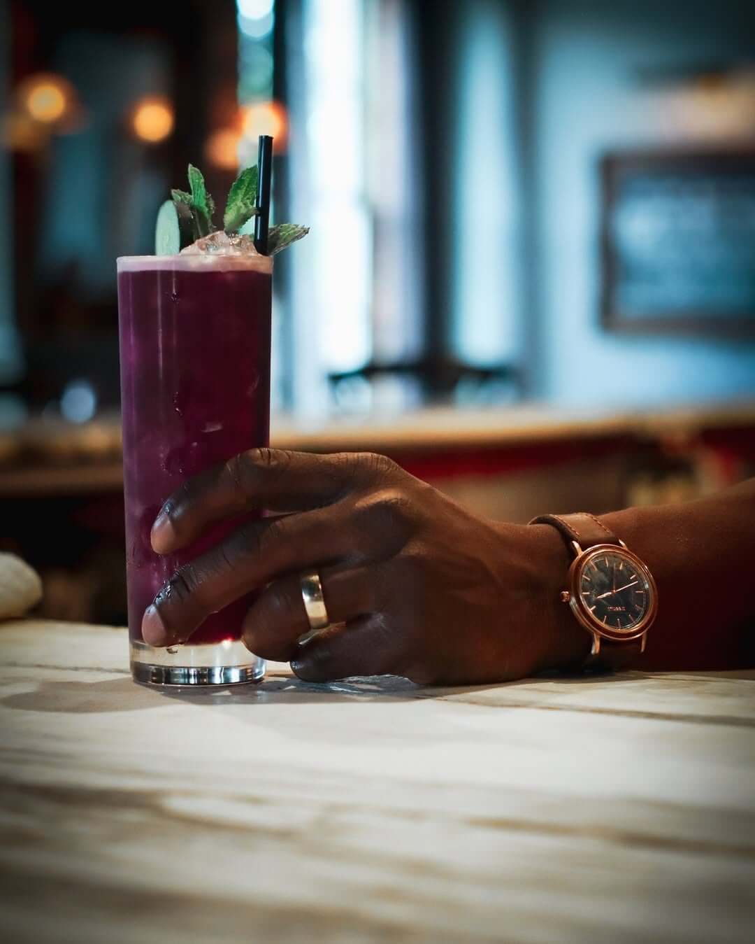 A hand wearing a watch and ring gracefully holds a tall glass of dark purple iced cocktail with mint garnish at The Marble Ring lounge in Avondale.