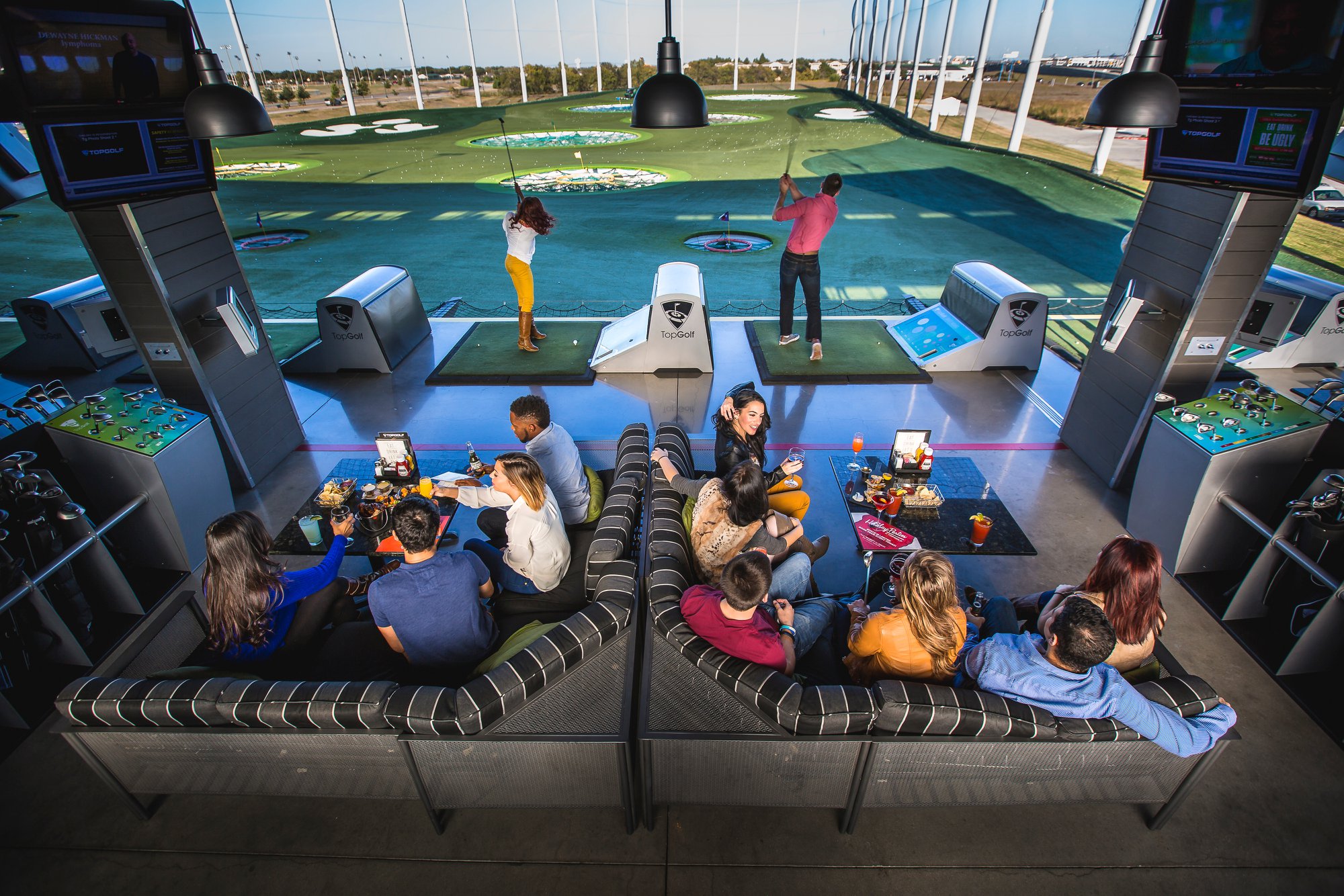 People are at an all-weather golfing bay at Topgolf. Several individuals unwind on couches with drinks and snacks while two golfers take their shots out onto the outfield. 
