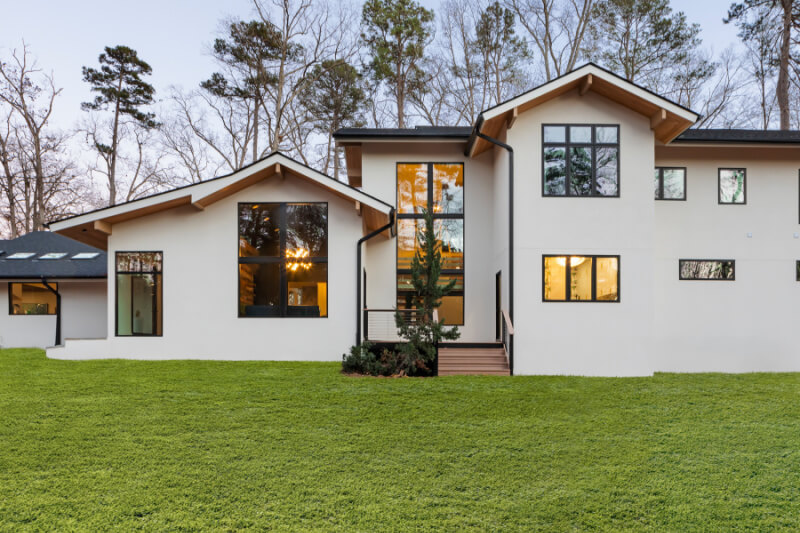 Modern two-story house with large windows, white exterior, and a sloped roof, surrounded by trees and a well-maintained lawn.