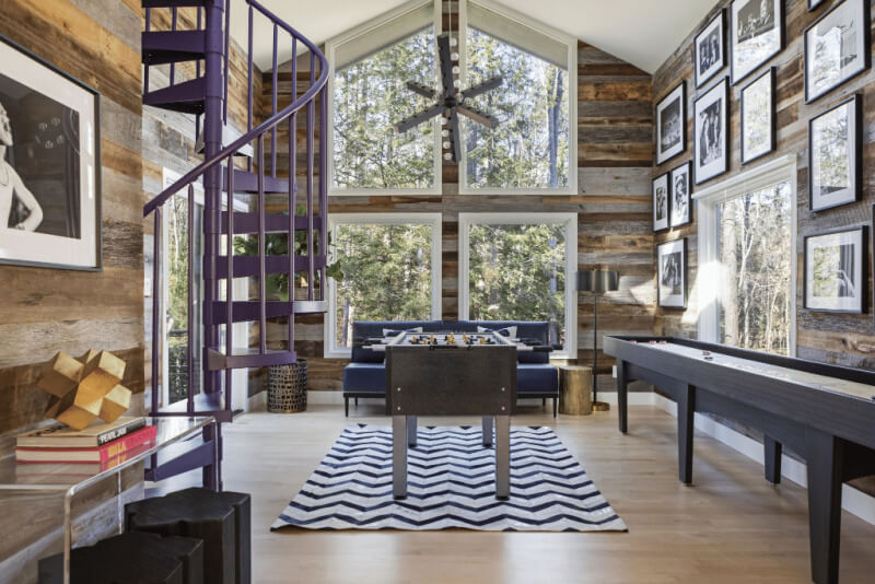 Rustic game room with purple spiral staircase, large windows, foosball table, shuffleboard, and photo-lined walls. Black and white rug on light wood floor.