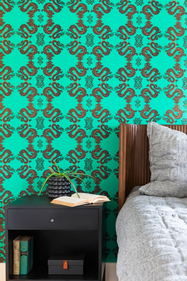 Bedroom with a wooden bed, gray bedding, and a black nightstand holding a book and decorative object. The wall features a vibrant green and brown patterned wallpaper.