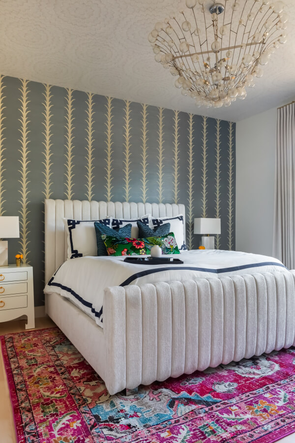 A bedroom with a white cushioned bed, colorful pillows, floral rug, and decorative wallpaper. A modern chandelier hangs from the ceiling. Two white nightstands have lamps and small decorations.