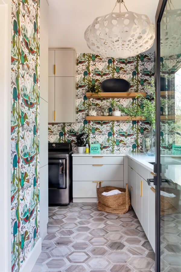 A small, modern kitchen with colorful wallpaper, white cabinets, open shelves, and a large, decorative hanging light. A wicker basket sits on a hexagonal-patterned floor.