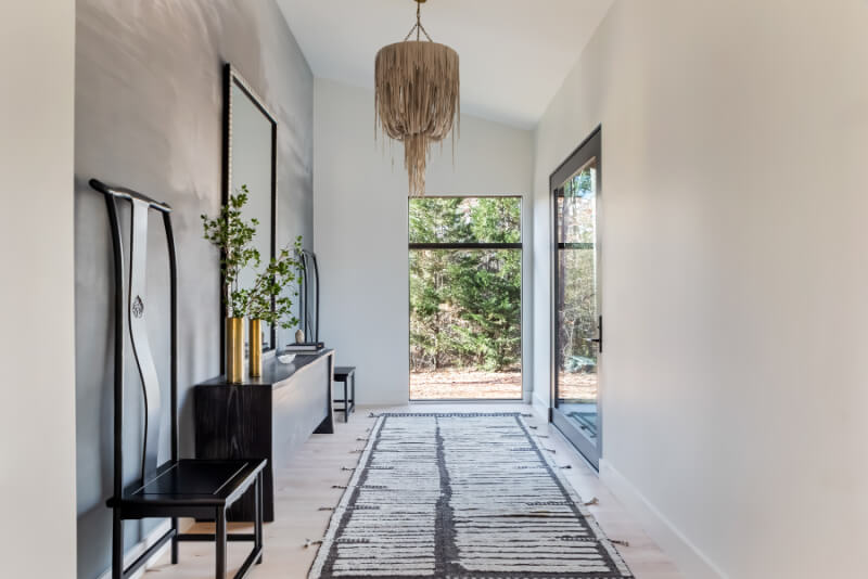 Modern hallway with a fringed chandelier, large mirror, two black chairs, a console table with green branches, and a patterned rug leading to a glass door with a view of trees outside.