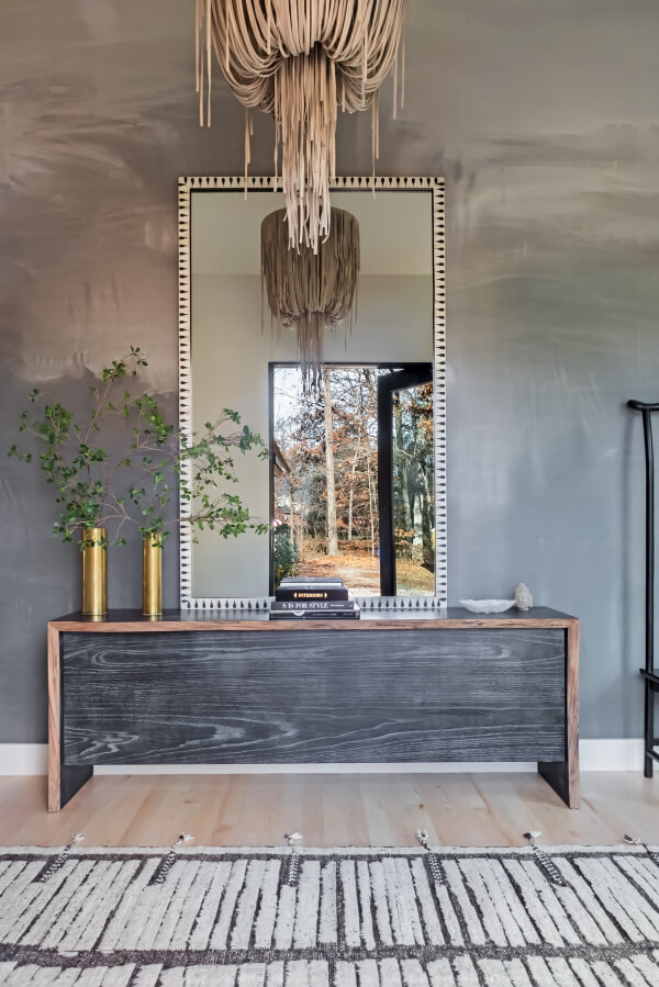 A stylish room with a large mirror reflecting an outdoor scene, a wooden console with plants and books, and a woven chandelier above. A geometric-patterned rug covers the floor.