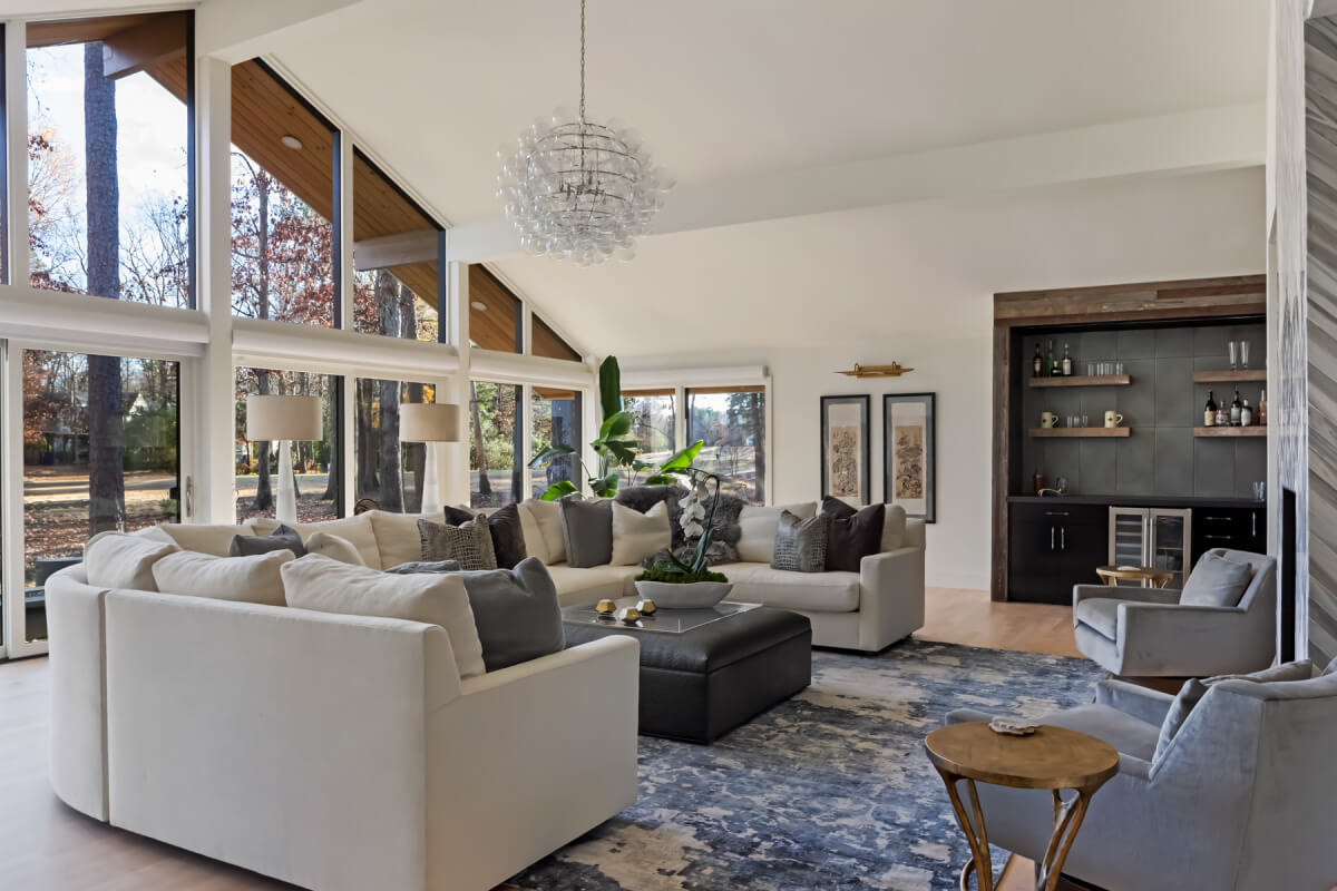 Spacious living room with large windows, white sectional sofas, a gray ottoman, and a modern chandelier. A bar area is visible in the corner.
