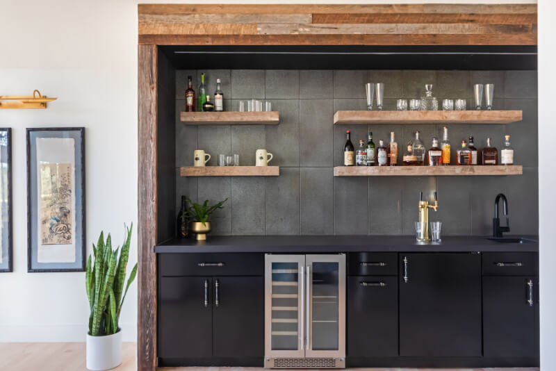Home bar with black cabinets, a small fridge, floating wooden shelves stocked with liquor bottles and glasses, a plant on the counter, and a snake plant beside the bar.