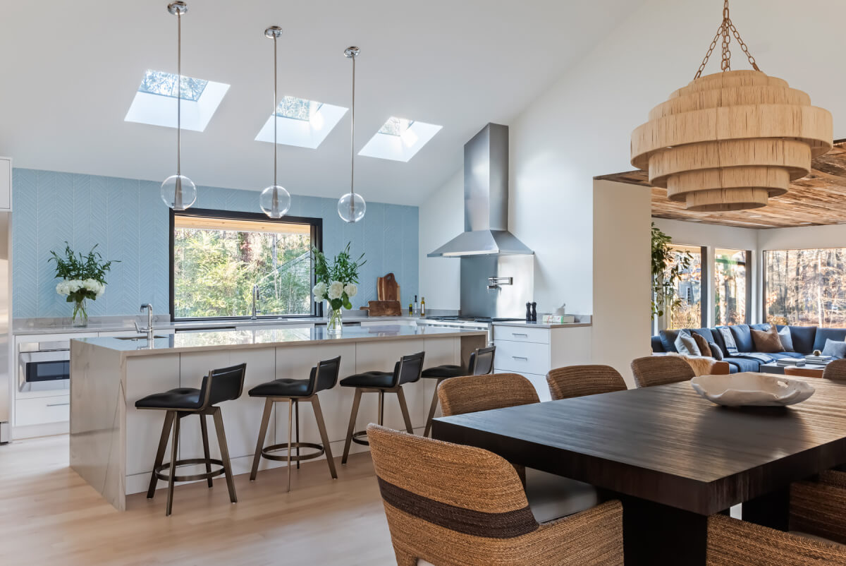 Modern kitchen and dining area with skylights. Features a large island with four stools, pendant lights, wooden table with chairs, and view into a living room through large windows.