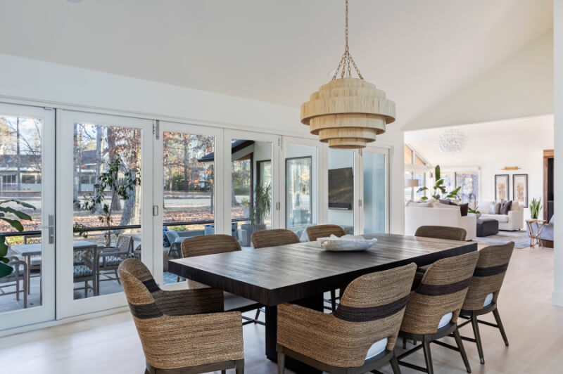 A modern dining room with a dark wood table surrounded by wicker chairs. Large windows and glass doors provide a bright, airy view of the outdoor patio area. A pendant light hangs above the table.