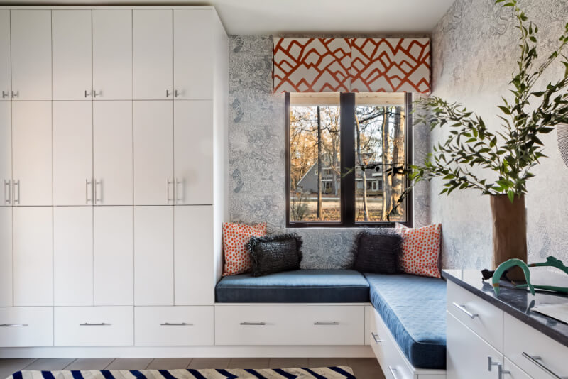 A cozy nook with cushioned seating and a striped rug next to white cabinets. A window with geometric-patterned valance overlooks a scenic view. Small plants add a touch of greenery.