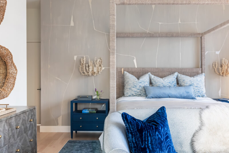 Stylish bedroom with a textured wall, a canopy bed, blue and patterned pillows, and a dark blue nightstand with decor. A woven mirror and a soft rug complete the modern look.