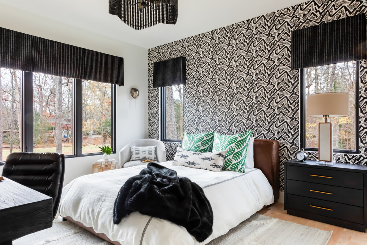 Bedroom with a bed, patterned accent wall, green and white pillows, black and white decor, windows with black Roman shades, a nightstand with a lamp, and an armchair by the window.