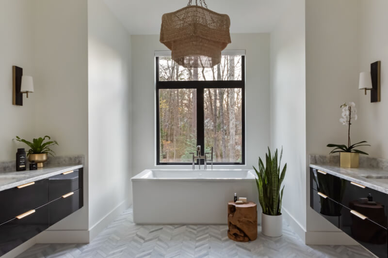 Modern bathroom with a central white bathtub beneath a large window, flanked by black cabinets. A potted plant and wooden stool are on the right, and a woven chandelier hangs above.