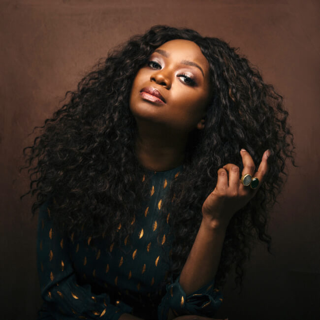 Ruby Amanfu, with her long curly hair, poses against a brown background. She wears a green patterned top and a ring.