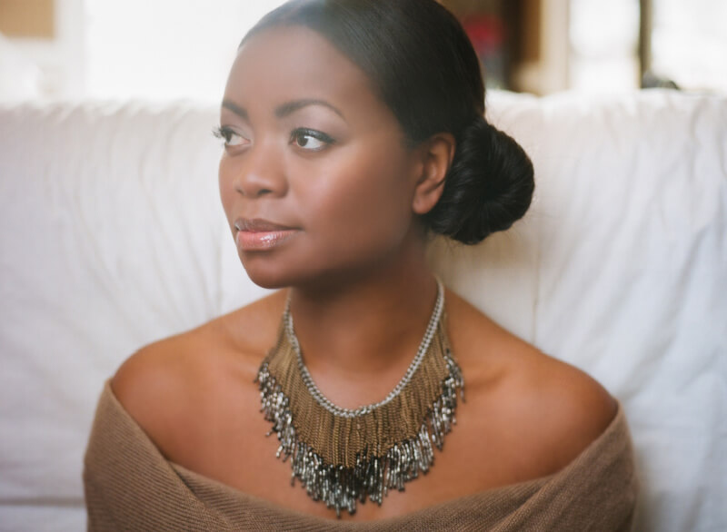 Ruby Amanfu, looking off-camera in a pensive stare. She is wearing an off-shoulder beige sweater and an ornate necklace.