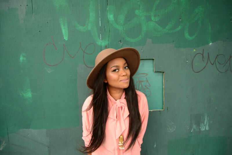 Ruby Amanfu wearing a brown hat and pink blouse stands against a green graffiti-covered wall.