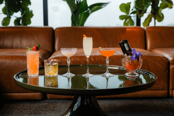 Five assorted cocktails in different glasses sit elegantly on a round table at PROOF Rooftop Bar, with a plush brown couch and lush green plants creating an inviting backdrop.