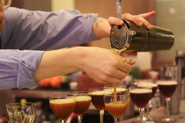 In March, at Mason Bar, the bartender deftly pours cocktails from a shaker into glasses, showcasing creations from the new beverage menu. Several completed drinks are perfectly lined up on the bar, ready to be enjoyed.
