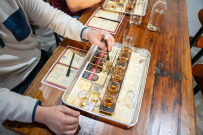 A person sits at a wooden table with a tray of various whiskey samples, tasting glasses, and a water carafe, exploring tasting menus with another participant—just like those offered in whiskey classes in Louisville.