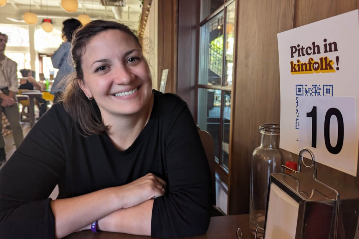 Emily Mutchnick sits at a table in a cozy cafe, her smile warm and inviting. In front of her is a sign displaying a QR code next to the number 10, capturing the blend of modern technology and classic charm that defines today's dining experience.