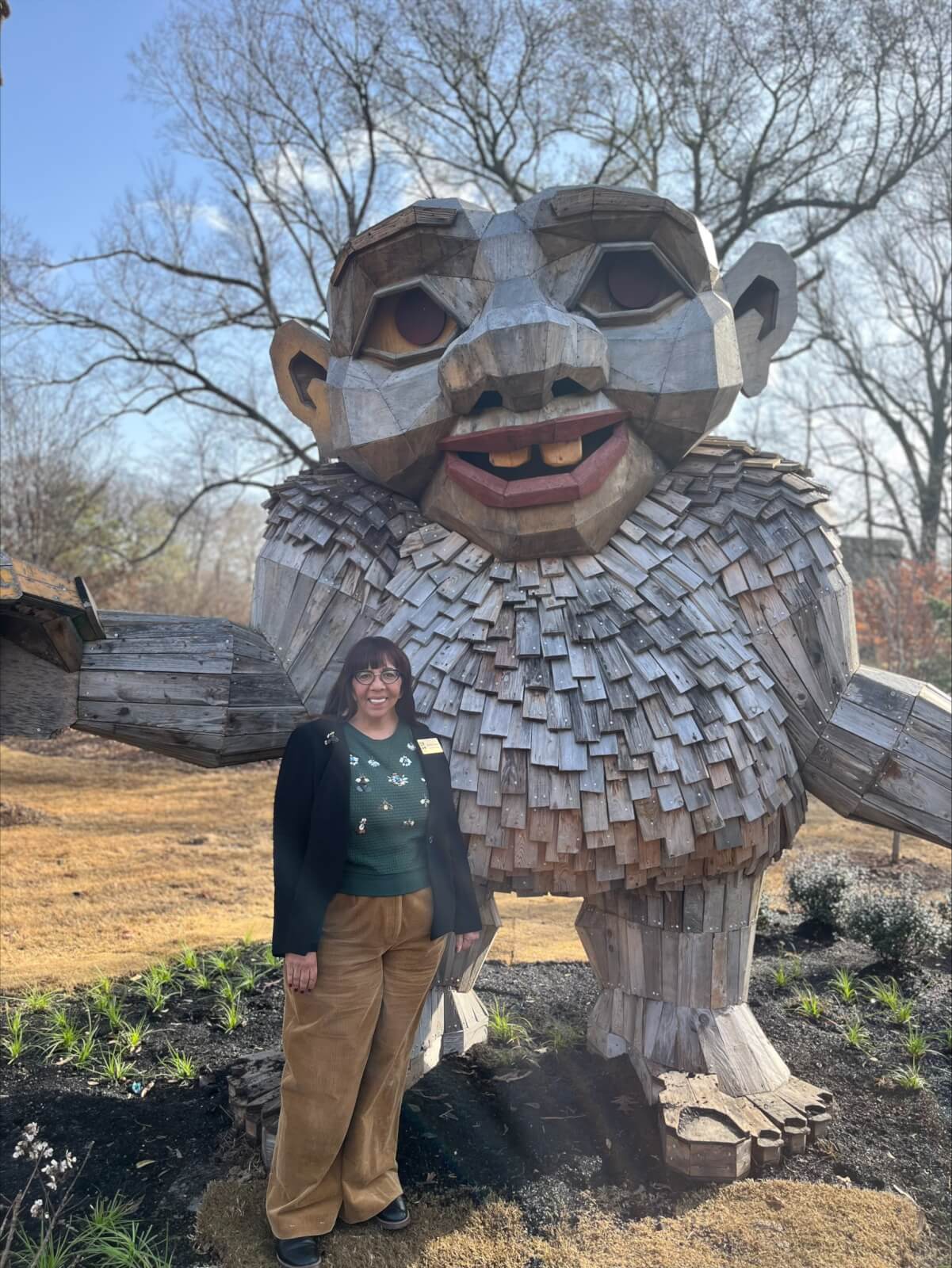 MaryLynn Mack stands next to a large wooden troll sculpture in an outdoor setting, surrounded by lush trees in the background.