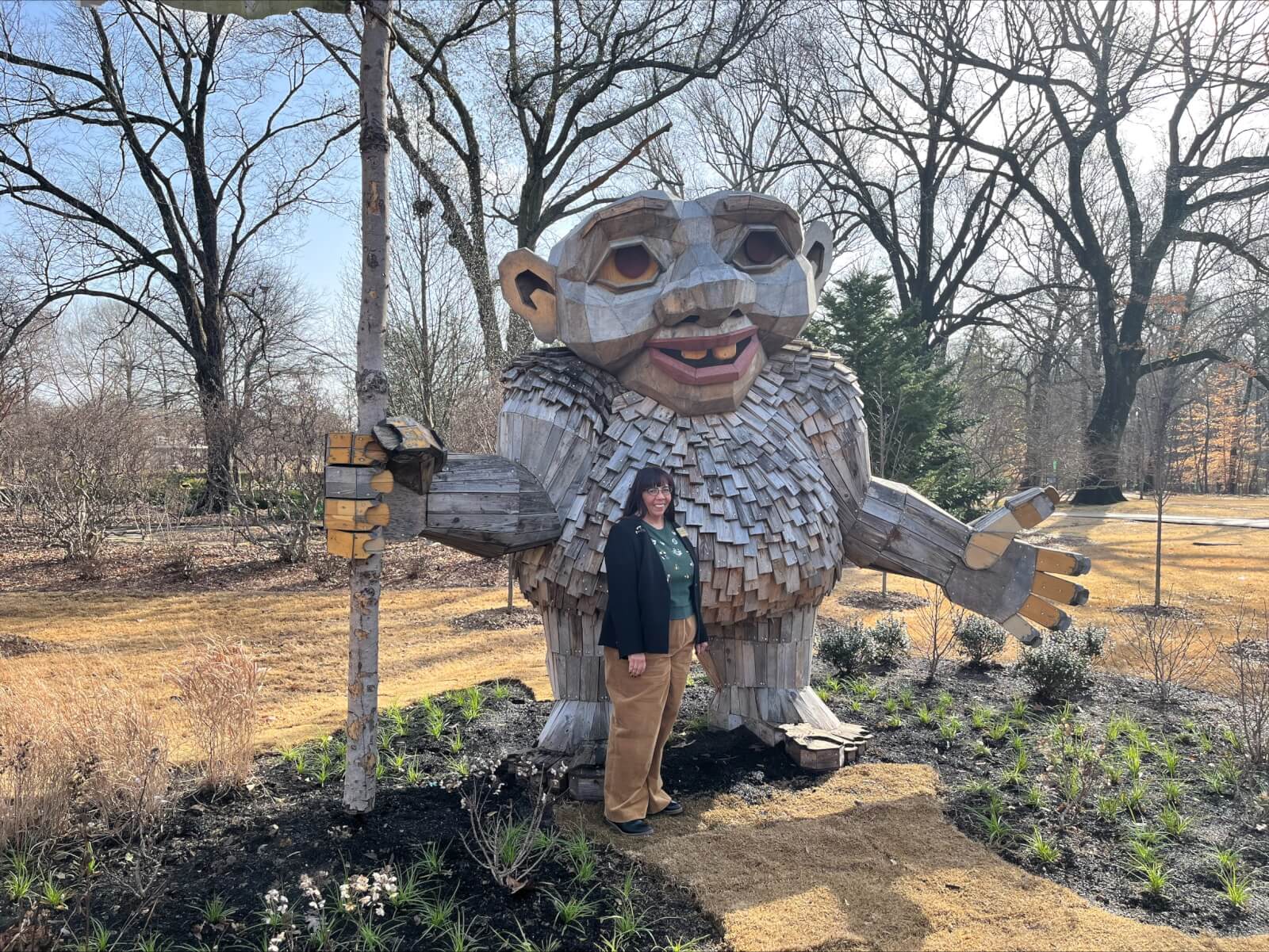 MaryLynn Mack stands next to a large wooden sculpture of a troll in an outdoor park, where towering trees create a lush backdrop.