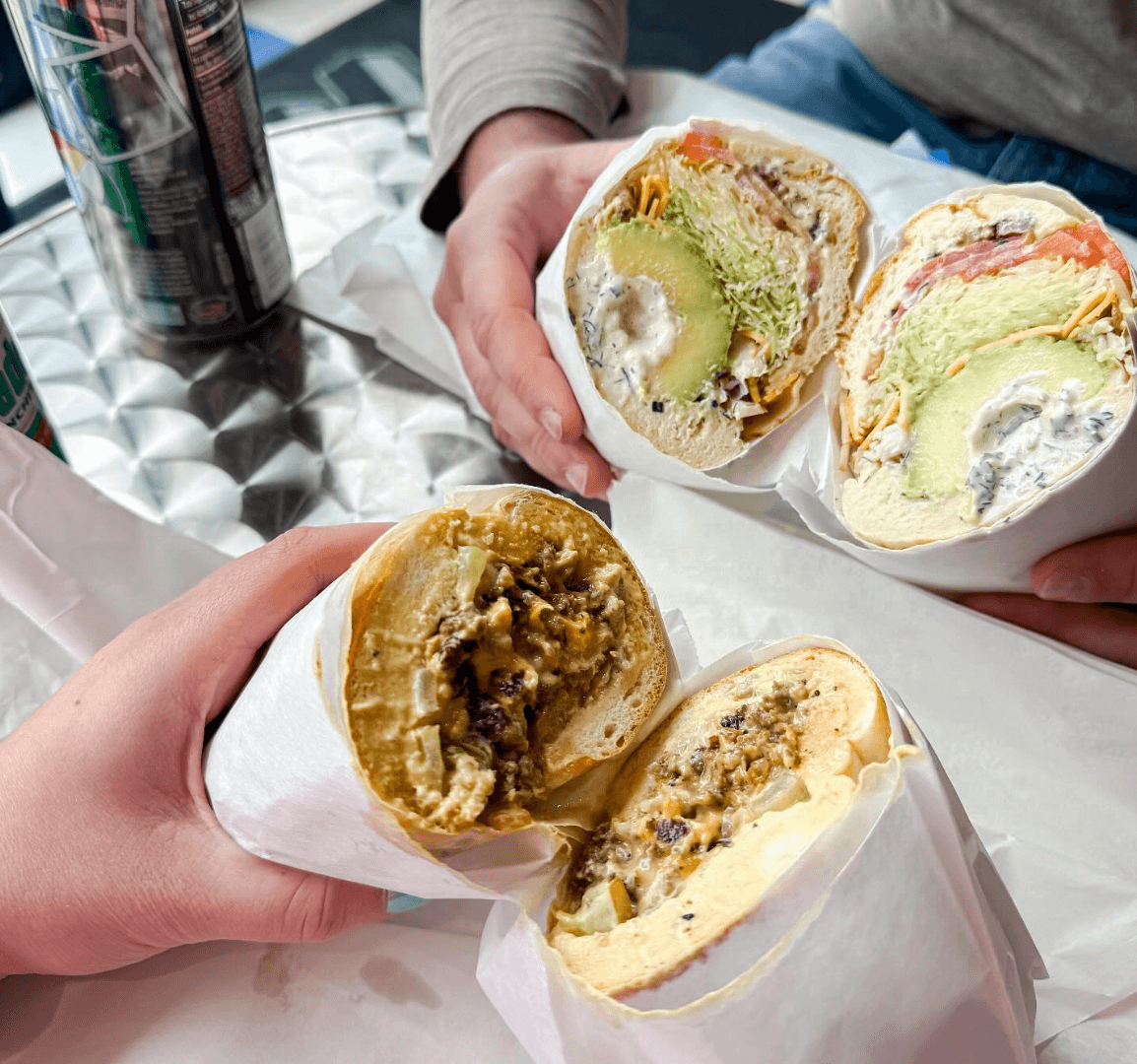 Two sandwiches wrapped in paper, held by different hands, brim with avocado and greens on a textured metal table. In the backdrop of this culinary delight are canned drinks, evoking the vibrant dining scene at new Memphis restaurants.