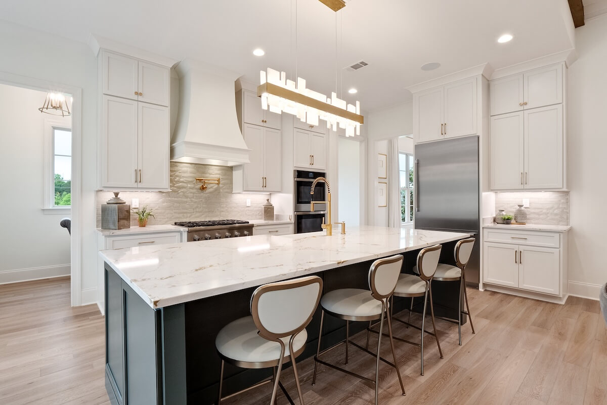 This Magnolia Homes modern kitchen features a large marble island with four chairs, stainless steel appliances, and sleek white cabinetry. A geometric light fixture gracefully hovers above, complementing the light wood flooring throughout.
