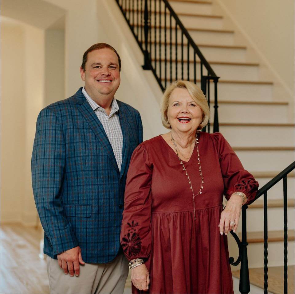 Two people standing together indoors in front of a staircase; the man in a blue jacket and the woman in a maroon dress.