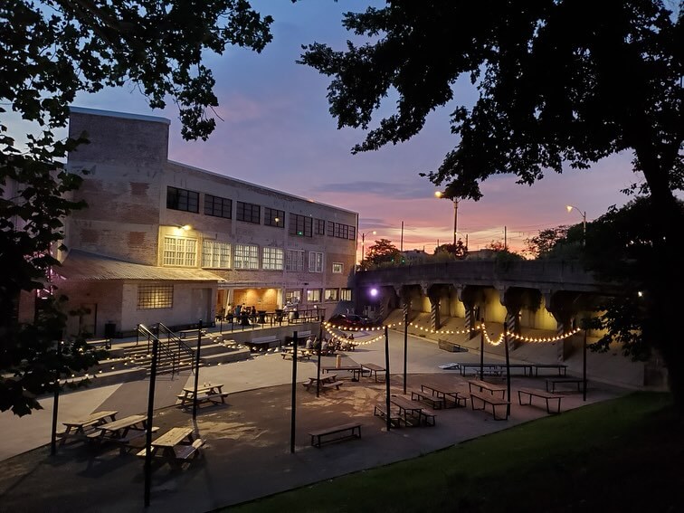 A courtyard in the Edge District features picnic tables and string lights, casting a warm glow at sunset beside an industrial-style building.
