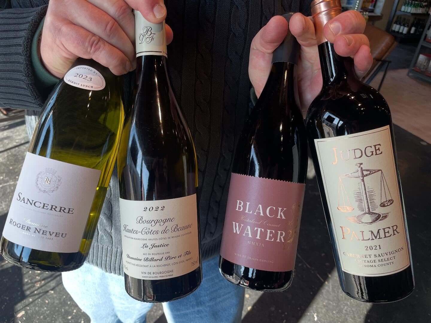 Four wine bottles held in hands: Sancerre 2023, Bourgogne 2022, Black Water Shiraz, and Judge Palmer Cabernet Sauvignon 2021—perfect selections for an Edge District tasting adventure.